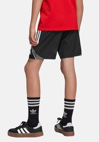 Zwarte sportshorts met drie witte strepen aan de zijkanten, gecombineerd met zwart-wit gestreepte sokken en zwarte sneakers. Rode shirt zichtbaar.