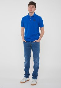 Polo shirt di cotone blu con toppa del marchio sul petto, abbinato a jeans azzurri e sneakers bianche con dettagli blu. Design semplice, maniche corte.