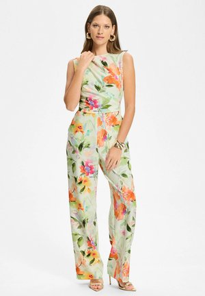 Femme portant une combinaison sans manches à imprimé floral en vert pastel avec des fleurs orange et roses, accessoirisée avec des boucles d'oreilles et un bracelet en or.