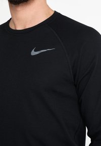 Schwarzes langärmliges Sportshirt aus strukturiertem Stoff. Mit einem grauen Nike-Logo auf der linken Brust und Raglanärmeln.