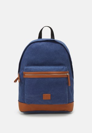 Sinine kangast seljakott, millel on pruun nahast põhi, lukuga esi-tasku ja must ülemine käepide. Sile tekstuur minimaalsete riistadega.