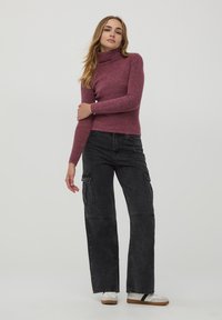 Maglione a collo alto bordeaux a costine abbinato a pantaloni cargo neri con tasche laterali. Il modello indossa sneaker bianche con suola marrone.