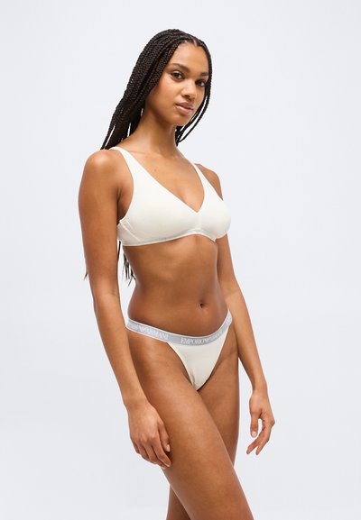 Γυναίκα με πλεξούδες που φορά λευκό τριγωνικό σουτιέν και ταιριαστό στρινγκ με το λογότυπο Emporio Armani στη μέση, στέκεται μπροστά σε μονόχρωμο φόντο.