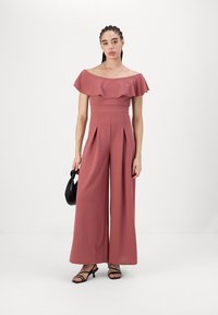 Jumpsuit v odstínu popelavé růže s odhalenými rameny, volánkovým výstřihem, plisé širokými nohavicemi a hladkou texturou, doplněný malou černou kabelkou.