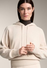 Žena so rovnými hnedými vlasmi a modrými očami, oblečená v krémovej mikine cez tmavý rebrovaný rolák, ktorá si komunikuje prstami.