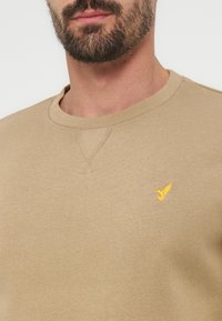 Άνδρας με κοντή γενειάδα που φοράει μπεζ φούτερ με στρογγυλή λαιμόκοψη και μικρό κίτρινο λογότυπο πουλί στο αριστερό στήθος.