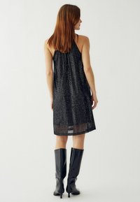 Čierne šaty s halter výstrihom, ktoré majú trblietavú textúru a voľný strih, doplnené vysokými lesklými čiernymi čižmami po kolená.