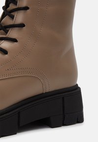 Botte de combat en cuir fauve avec semelle épaisse noire et lacets noirs, présentant des coutures détaillées et un motif de bande de roulement texturé.