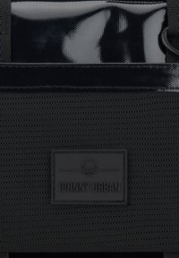 Zaino nero con un tessuto testurizzato e una parte superiore lucida e liscia. Include un'etichetta con logo visibile e testo in rilievo.