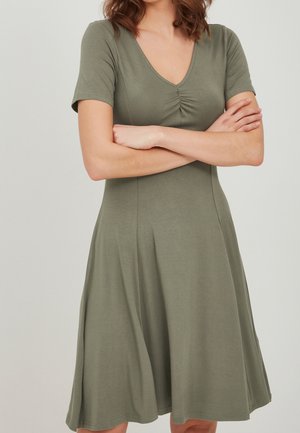 Kvinde iført en kortærmet olivengrøn kjole med V-hals og sammensat detalje, stående med armene over kors mod en ensfarvet baggrund.