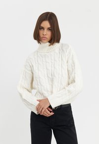 Pull en tricot blanc chunky avec un col haut à côtes et des manches larges, associé à un jean noir. Texture douce et coupe décontractée.