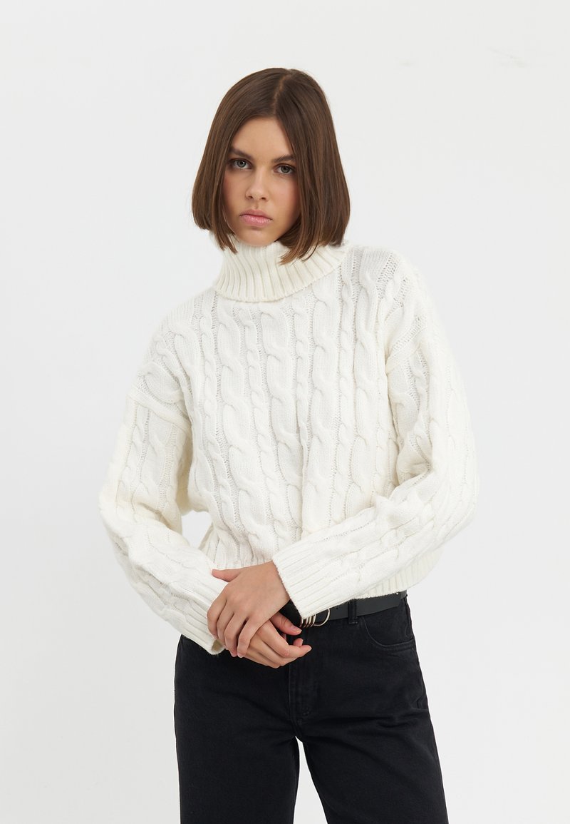 Pull en tricot blanc chunky avec un col haut à côtes et des manches larges, associé à un jean noir. Texture douce et coupe décontractée.