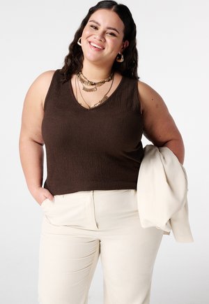 Vrolijke vrouw met donker haar draagt een mouwloos bruin shirt, meerdere gouden kettingen, crèmekleurige broek en houdt een lichtgekleurde jas over haar arm.