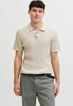 Fiatal férfi, aki krémszínű, kötött rövid ujjú galléros ingben és szürke nadrágban, fekete fonott övvel áll egyszínű háttér előtt.