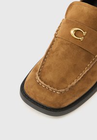 Καφέ μοκασίνι από σουέντ με ραμμένο σχέδιο, στρογγυλεμένη μύτη και χρυσή λογότυπη λεπτομέρεια. Πάτος παχύς μαύρος με υφή στην άκρη για καλύτ ερη πρόσφυση.