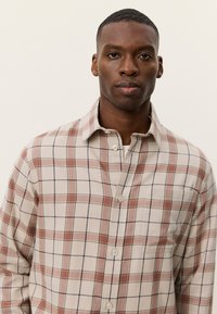 Καρό πουκάμισο σε ανοιχτό μπεζ με κόκκινα, πράσινα και μπλε σχέδια καρό· διαθέτει μπροστινά κουμπιά, γιακά και μία τσέπη στο στήθος.