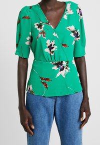 Blusa verde floral con escote en V, mangas cortas abullonadas y cintura ajustada. Fabricada con un tejido ligero de acabado texturizado.