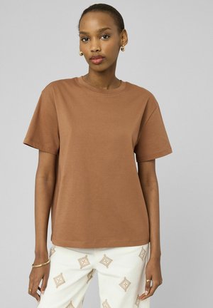 Femme portant un t-shirt marron à manches courtes et un pantalon blanc avec des motifs géométriques beiges, debout sur un fond gris clair.