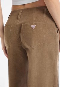Hnědé kordové kalhoty se zadními kapsami, přičemž jedna má malou trojúhelníkovou nášivku s logem Guess, oblečené osobou z pohledu od pasu dolů.