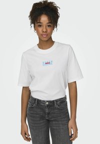 Witte katoenen T-shirt met korte mouwen met blauwe en rode tekst "BEACH ESSENCE BRAZIL" afgedrukt op de voorkant. Draagt het met donkere spijkerbroeken.
