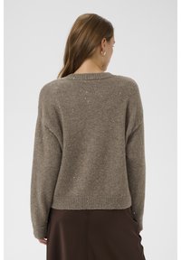 Moteris su ilgais rudos spalvos plaukais, dėvinti auksinius lankinukus, rudą margą megztinį ir tamsiai rudą sijoną, stovinti nugara.