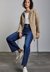 Beige huva jacka med dragsko, bärs över en vit skjorta. Mörkblå vida jeans och vita sneakers med beige detaljer. Sittande hållning.