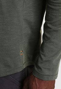 Olijfgroene, langsteek shirt met een ronde hals, textuurstof en oranje stiksel detail op de zoom.
