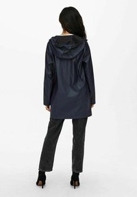 Imperméable marine à capuche, au design simple et à la texture lisse. Assorti à un pantalon large gris foncé et des bottes à talons noires.