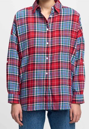 Person wearing a loose red, blue, and green plaid button-up shirt with rolled-up sleeves and blue jeans, standing against a white background.