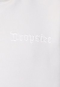 Bijela tkanina s reljefnom bijelom izvezom riječi "Dropsize" u stiliziranom, gotičkom fontu, stvarajući teksturirani izgled.