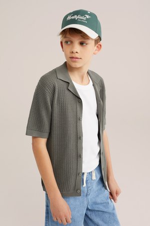 Berniukas dėvi žalią Northfield Falcons kepurę, pilką trumpomis rankovėmis marškinius su sagomis ant baltos marškinėlių ir šviesiai mėlynas džinsus, žiūri į šoną.