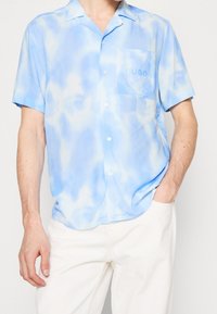 Ljusblå kortärmad skjorta med ett vatt färgat tie-dye mönster, med en knappstängning fram och en vänster bröstficka som visar logotypen "HUGO."