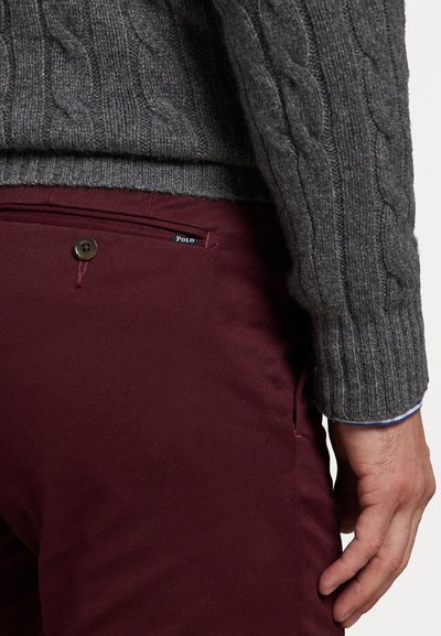 Šedý svetr s pleteným vzorem a burgundské kalhoty. Svetr má žebrovaný výstřih a manžety. Kalhoty obsahují kapsu na knoflík s logem.