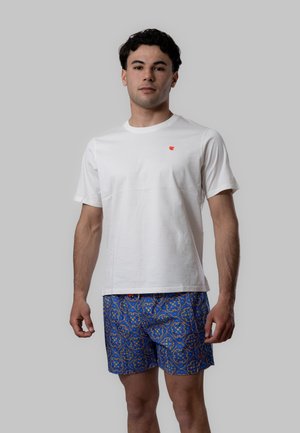 Giovane uomo in piedi, indossa una maglietta bianca semplice con un piccolo logo rosso e pantaloncini blu a motivi con dettagli gialli e arancioni.