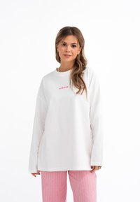 Felpa bianca a maniche lunghe con collo rotondo che presenta la scritta "MIRONS" in rosa. Tessuto morbido, vestibilità oversize. Modello in piedi con pantaloni a righe rosa.