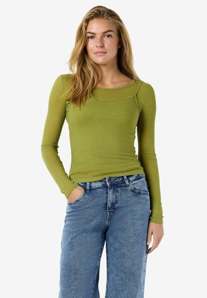 Vrouw met lang blond haar, gekleed in een olijfgroen shirt met lange mouwen en losse blauwe spijkerbroek, staand met één hand in de zak.