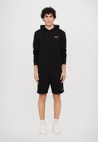 Black hoodie with adjustable drawstrings and front pocket, paired with black shorts featuring a small gold logo. White sneakers complete the look.