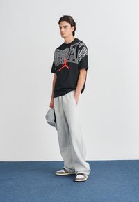 Μαύρη oversized μπλούζα με γραφικά "Jordan" και κόκκινο λογότυπο, συνδυασμένη με ανοιχτό γκρι παντελόνι φόρμας. Φόρεμα έως το πάτωμα, casual, με μαλακή υφή.