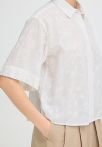 Blouse blanche à manches courtes avec une broderie florale subtile, portée rentrée dans un pantalon beige plissé, main posée négligemment dans la poche.