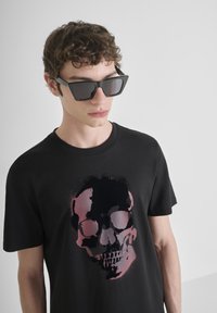Camiseta de algodón negra con un gran gráfico de calavera en colores rosa y negro. El modelo lleva gafas de sol negras y angulares.
