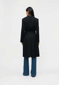 Γυναίκα που στέκεται με την πλάτη, φοράει μαύρο παλτό μέχρι το γόνατο πάνω από μπλε φαρδιά τζιν παντελόνια, μπροστά σε απλό γκρι φόντο.