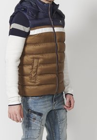 Brown padded vest with a navy blue, hooded top section. Features horizontal white stripe, front zipper, and side pocket. Textured sleeves visible.