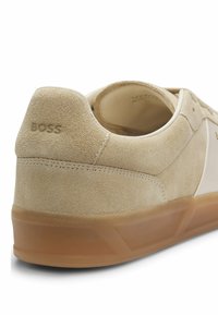 Beige ruskindssneaker med gummisål, synlige syninger og "BOSS"-logo præget på hælkappen.