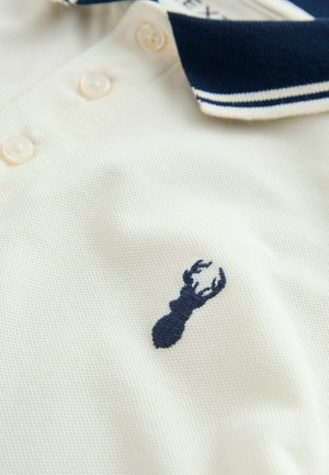 Polo majica u krem boji s mornarsko plavim ovratnikom i prugama. Ima mornarsko plavi vezeni motiv jastoga na prsima. Izrađena od teksturiranog tkanja.