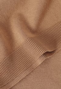 Tissu tricoté beige avec une texture lisse et un détail de bord-côte côtelé. Le matériau doux présente des variations subtiles de tissage et de profondeur de couleur.