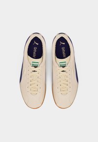 Ζευγάρι μπεζ αθλητικά παπούτσια Puma με εκρού κορδόνια, σκούρες μπλε εσωτερικές σόλες με ετικέτα Ortholite και πράσινο λογότυπο Puma στη γλώσσα, εμφανιζόμενο από πάνω.