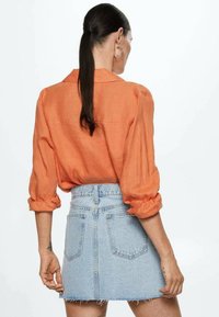 Camisa de lino naranja con las mangas remangadas, combinada con una minifalda de mezclilla azul claro, que presenta un dobladillo desflecado y bolsillos traseros.