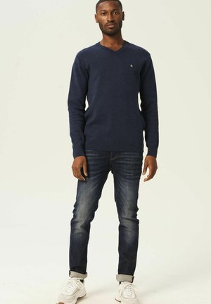 Pull en V bleu marine en matériau doux, associé à un jean foncé et des baskets blanches, avec un petit logo sur la poitrine.