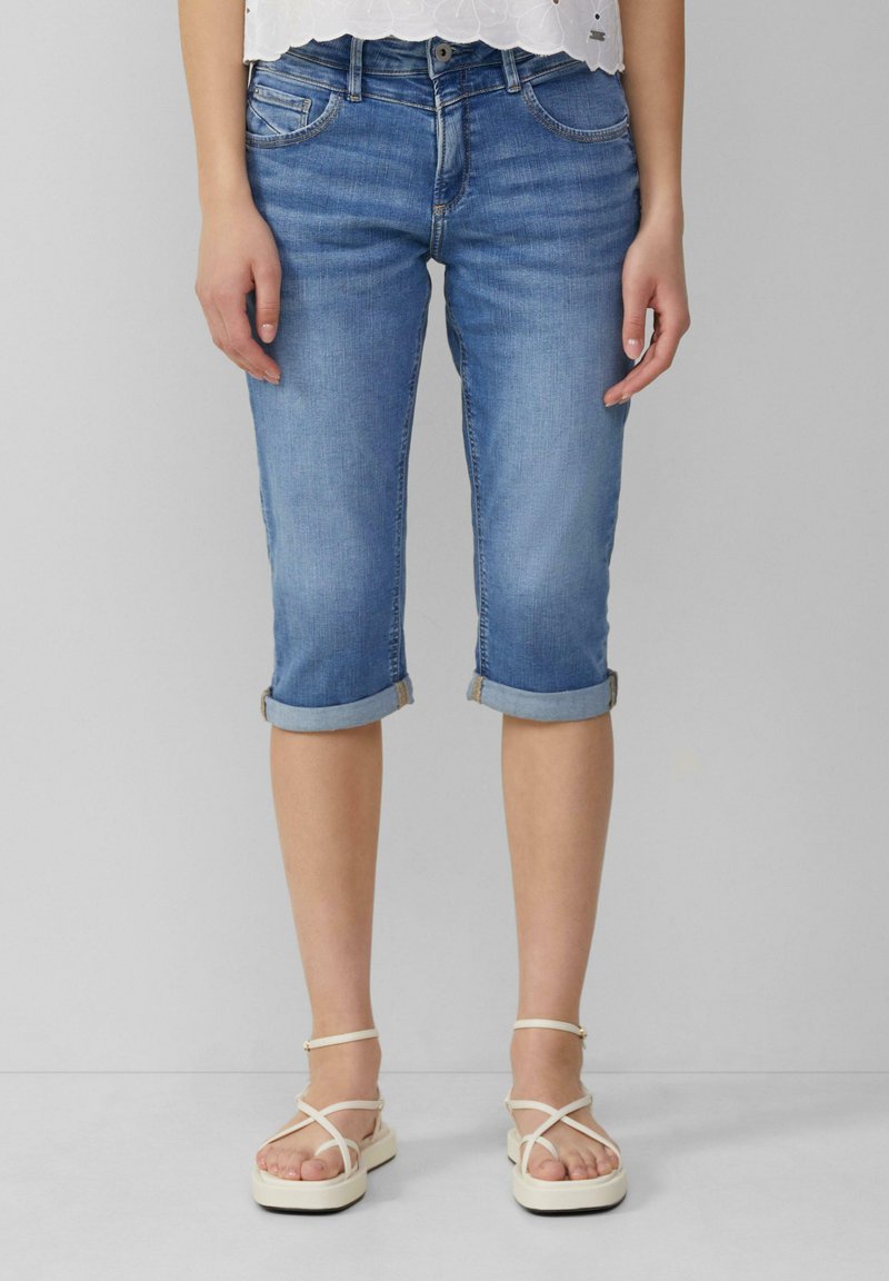 Jean capri bleu clair avec des ourlets roulés, style cinq poches et un léger délavage. Associés à des sandales à lanières crèmes.