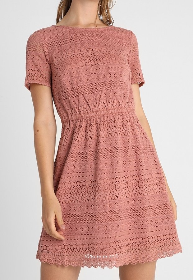 Femme portant une robe en dentelle rose poudrée à manches courtes avec des motifs floraux et géométriques et une taille cintrée.